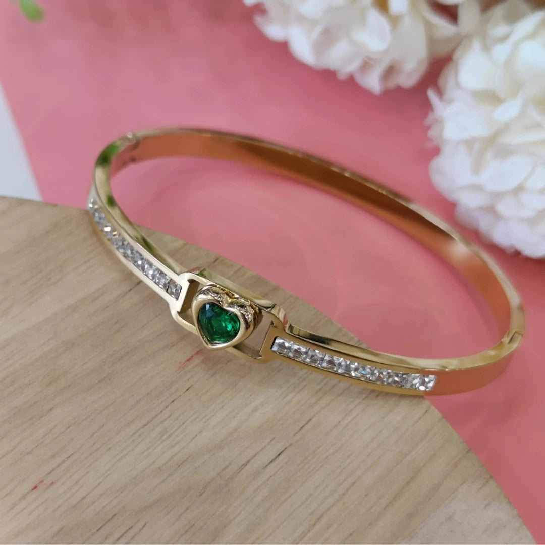 Jwanas Emerald-Green Heart Bangle Bracelet resting on a wooden surface with white flowers and a pink background
