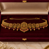 Jwanas Royal Lakshmi Temple Gold Kamarbandh featuring an ornate goddess centerpiece and ruby-red accents, displayed in a burgundy velvet jewellery box.